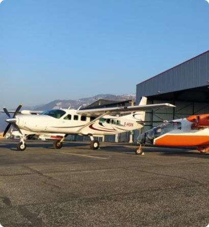 Deux petits avions étiquetés « sintegra » sont stationnés sur le tarmac dun aéroport, devant limpressionnant Hangar de SINTEGRA. Lun des avions est blanc, tandis que lautre arbore un design orange et blanc saisissant. Un ciel clair et des collines lointaines encadrent le fond serein.