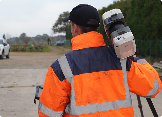 Un géomètre expert, vêtu d'une veste et d'une casquette orange réfléchissantes, se tient dehors, un trépied sur l'épaule et une tablette à la main. En arrière plan, un sentier, de la verdure et une voiture blanche, mettent en valeur les outils de précision de son métier.